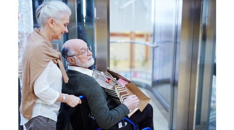 Ar senjorams kompensuos sanatorinį gydymą? Nuotraukoje žilas senjoras sėdi neįgaliojo vežimėlyje šalia jo stovi senjorė šypsodamasi
