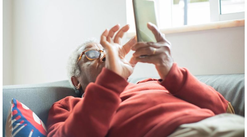 Kokios pensijos ir išmokos priklauso senjorams Nuotraukoje senjoras guli ant lovos ir iškeltoje rankoje laikydamas mobilųjį telefoną kita ranka naršo.